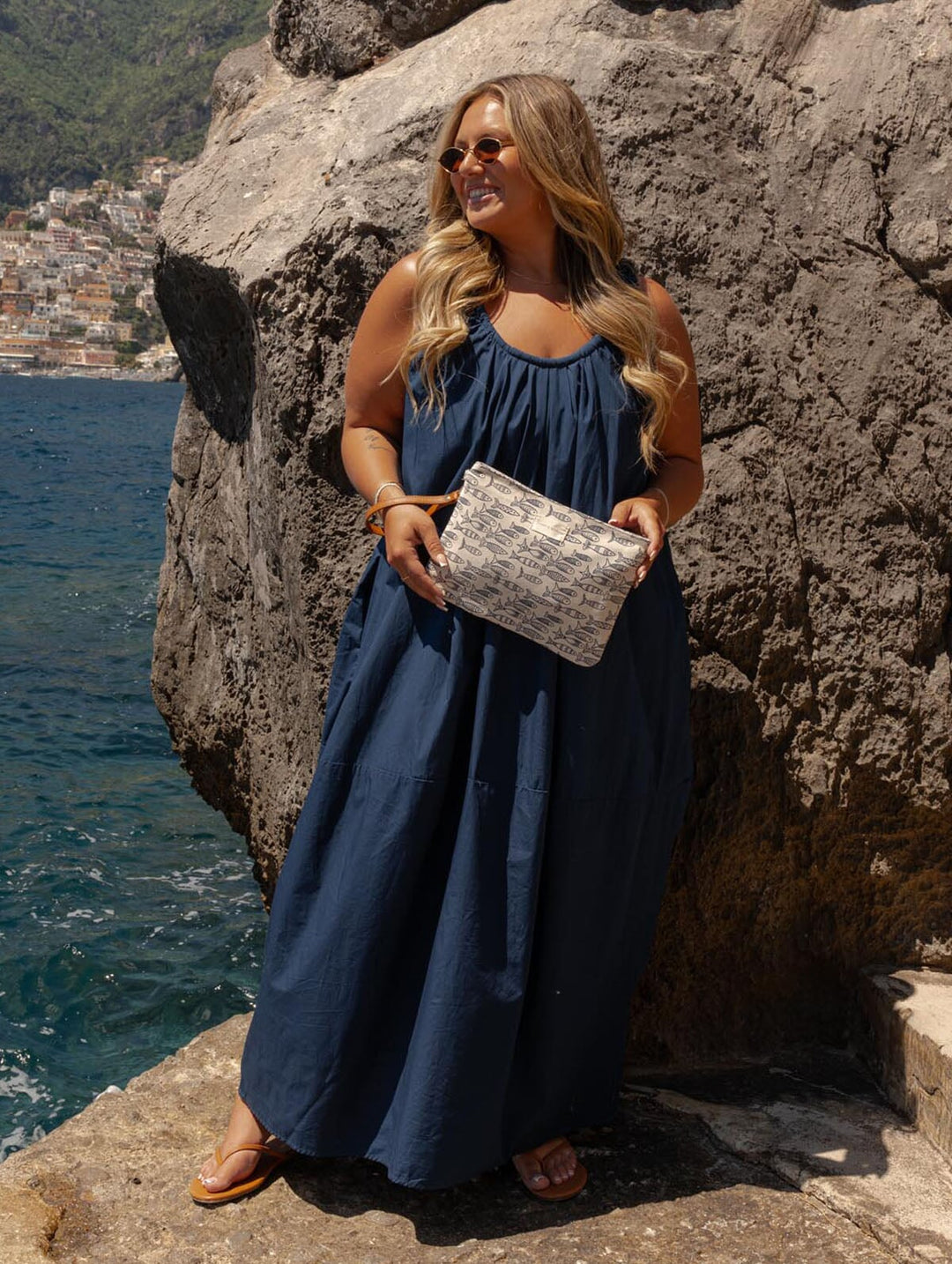 Woman in a blue dress holding a sardine patterned against a rocky cliff with ocean view