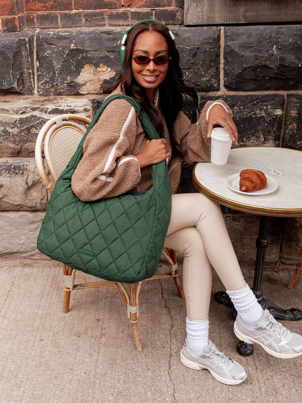 Woman sitting at a table with a green quilted bag, wearing sunglasses and holding a coffee cup.