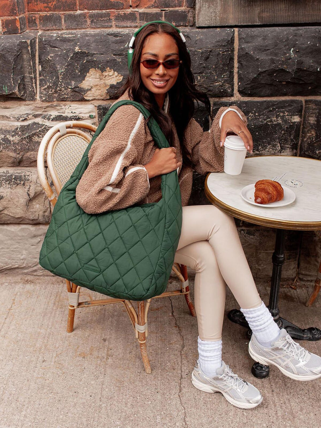 Woman sitting at a table with a green quilted bag, wearing sunglasses and holding a coffee cup.