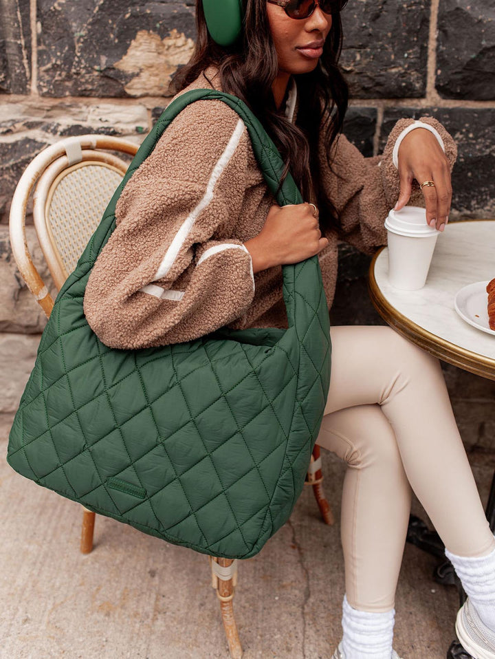 Person sitting at an outdoor cafe with a green quilted bag and a cup of coffee.