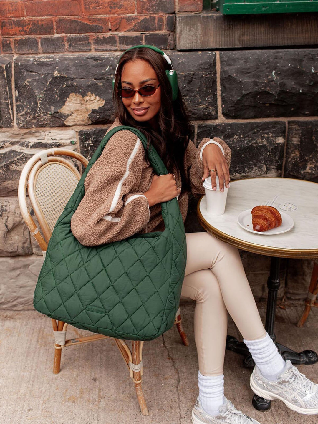 Woman sitting at a cafe table with a green quilted bag, wearing headphones and sunglasses.