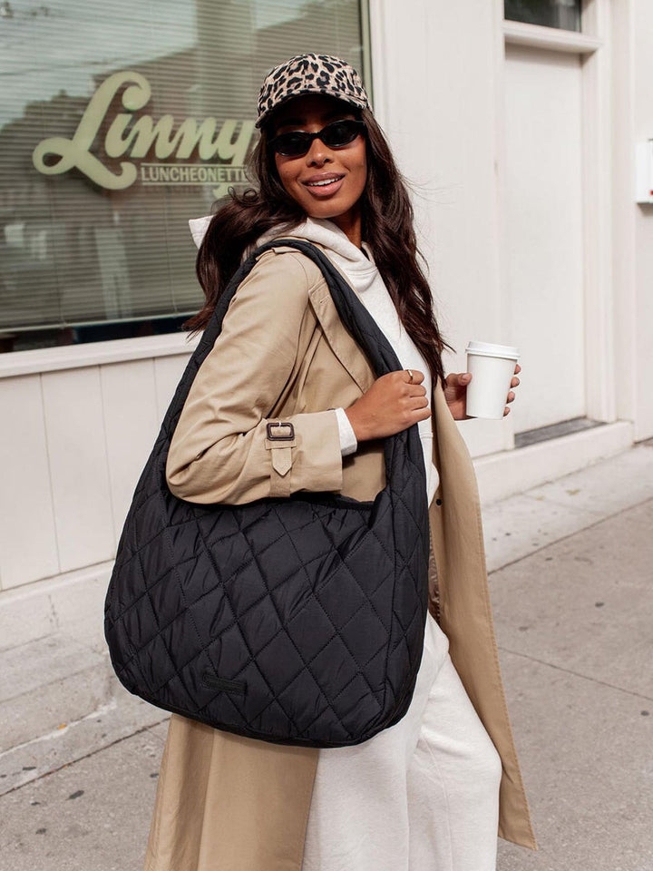 Woman holding a black quilted bag and a coffee cup, wearing a leopard print cap and sunglasses, standing on a sidewalk.