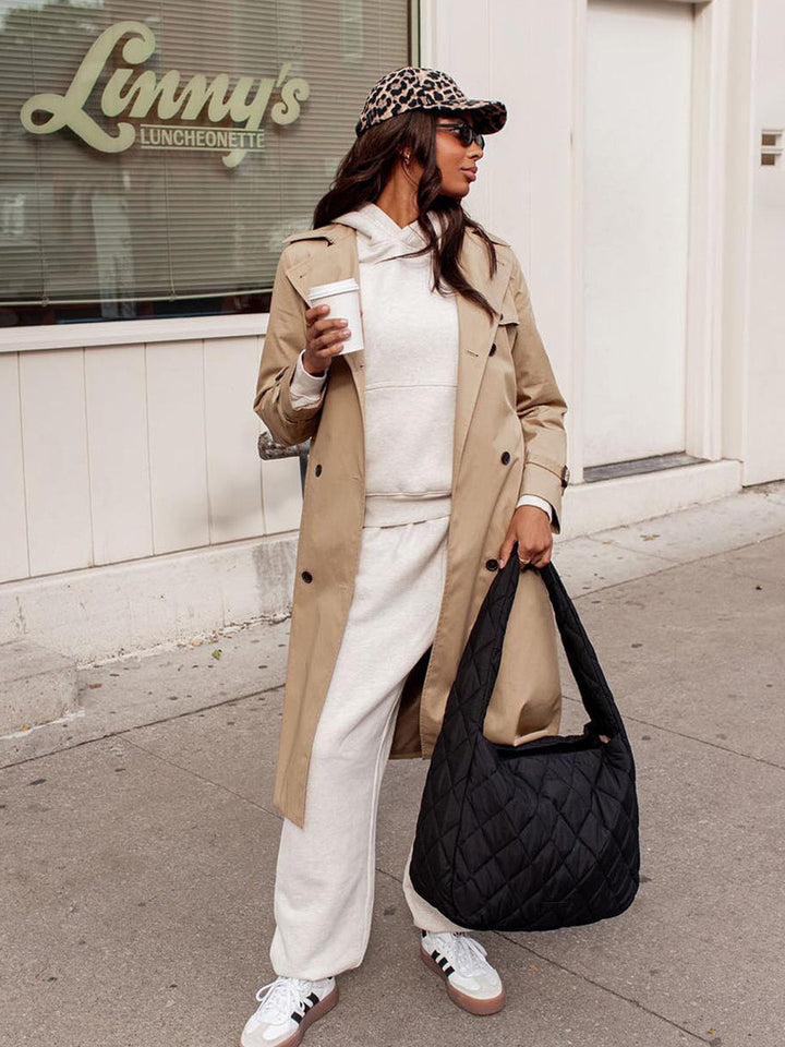 Woman in trench coat holding a coffee cup and black quilted bag on a sidewalk.