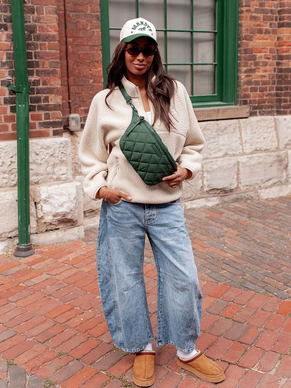 Person wearing a cream sweater, blue jeans, and brown shoes, holding a green quilted bag on a brick street.