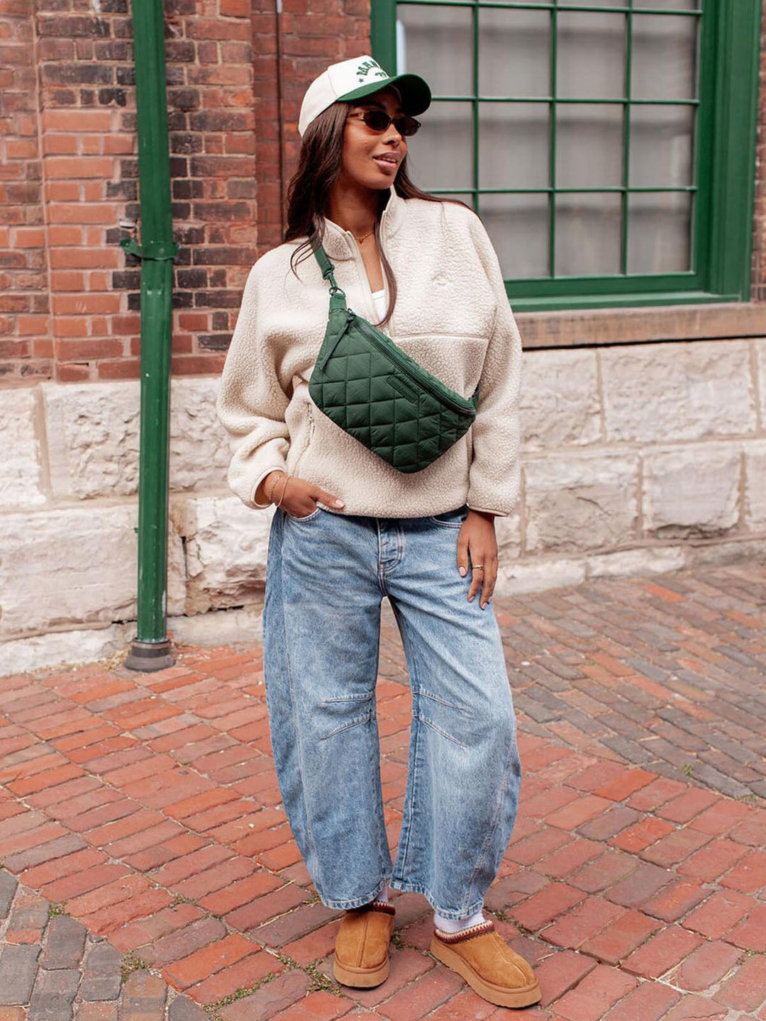 Woman wearing a cream sweater, blue jeans, tan shoes, and a green quilted bag on a brick sidewalk.