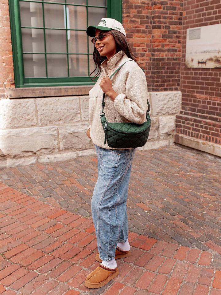 Woman in a beige sweater and jeans standing on a brick sidewalk with a quilted green bag.