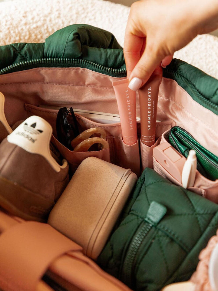 Hand reaching into a green quilted tote bag with various items including a pink tube of lip balm.