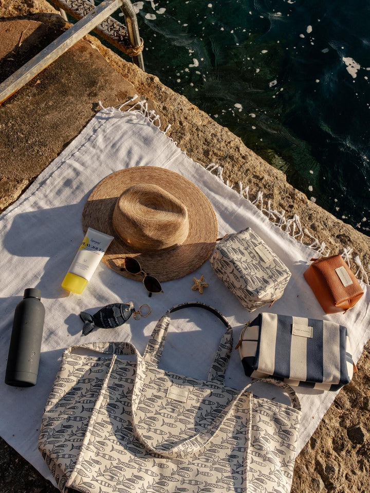Sun hat, bottle, and printed tote bag on a towel by the water