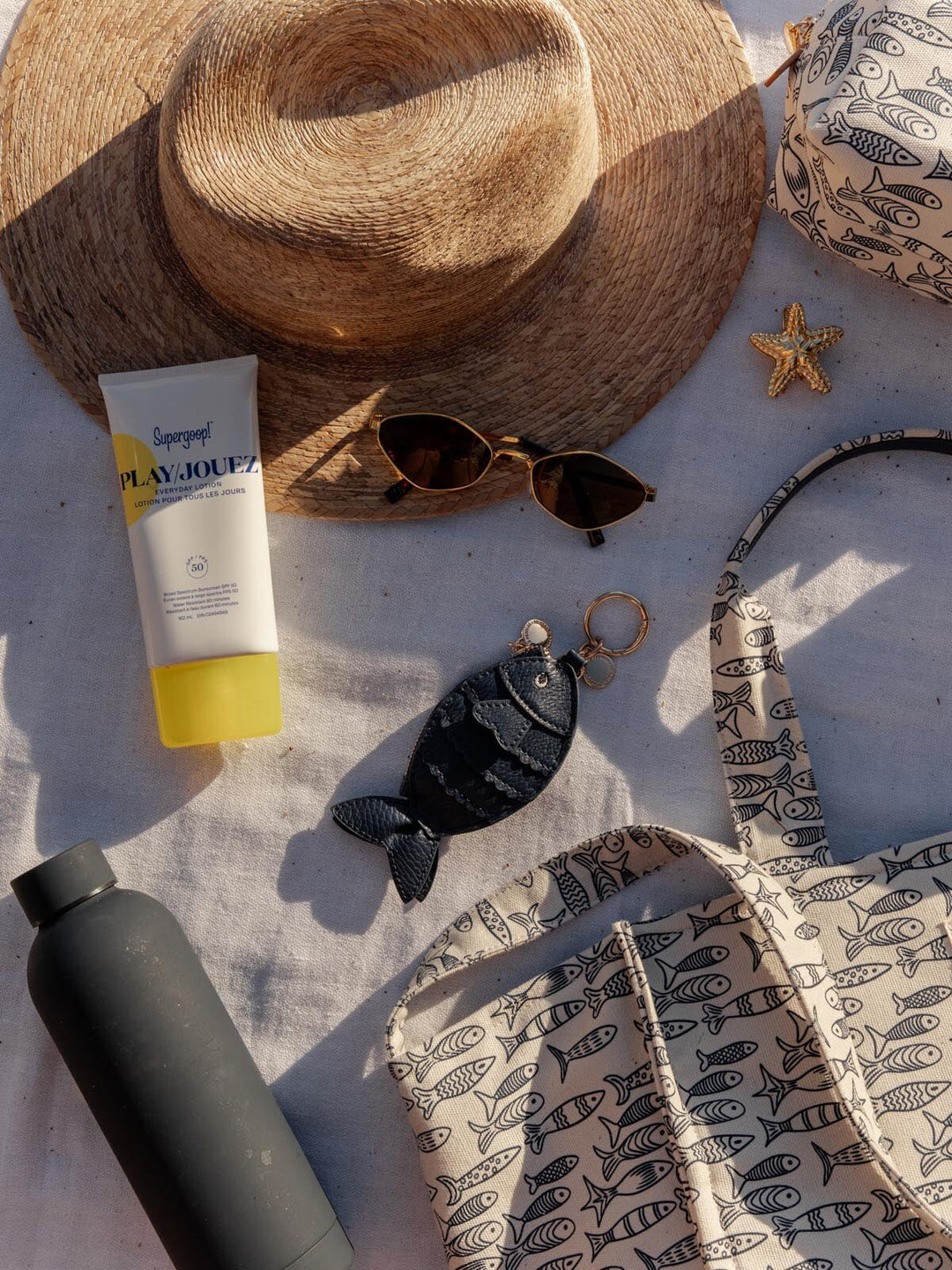 Fish bag charm, sunscreen bottle, sunglasses, straw hat, and patterned bag on a textured surface