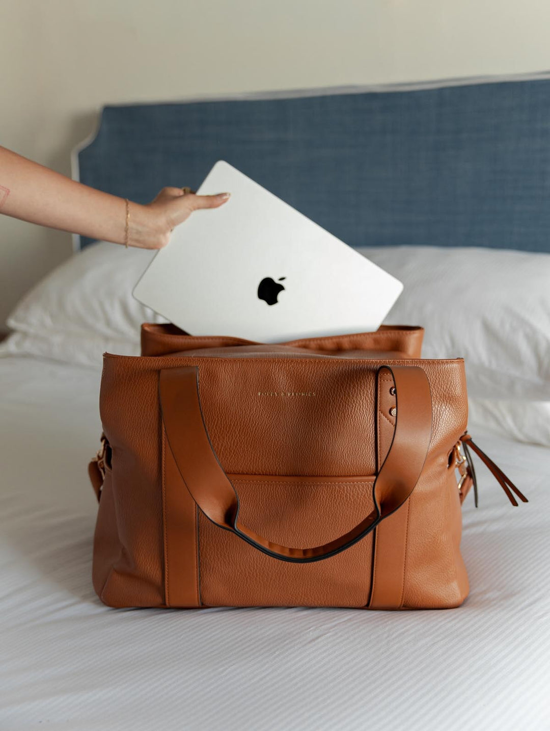 Brown vegan leather travel bag on a bed with an Apple laptop being placed inside