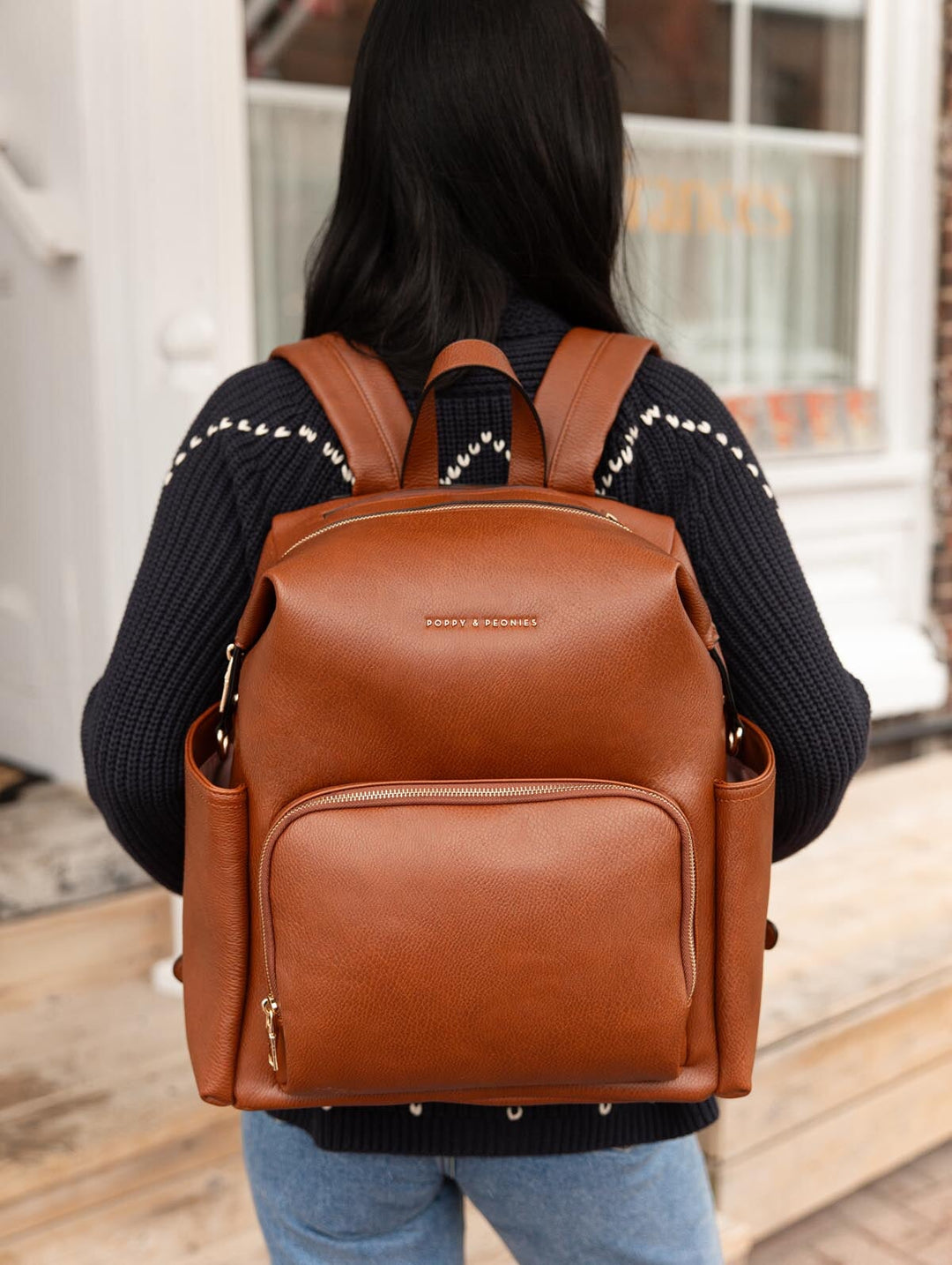 Poppy & Peonies The Cabin Backpack in Chestnut worn on the back by a woman in a navy cardigan and blue jeans outside a storefront.
