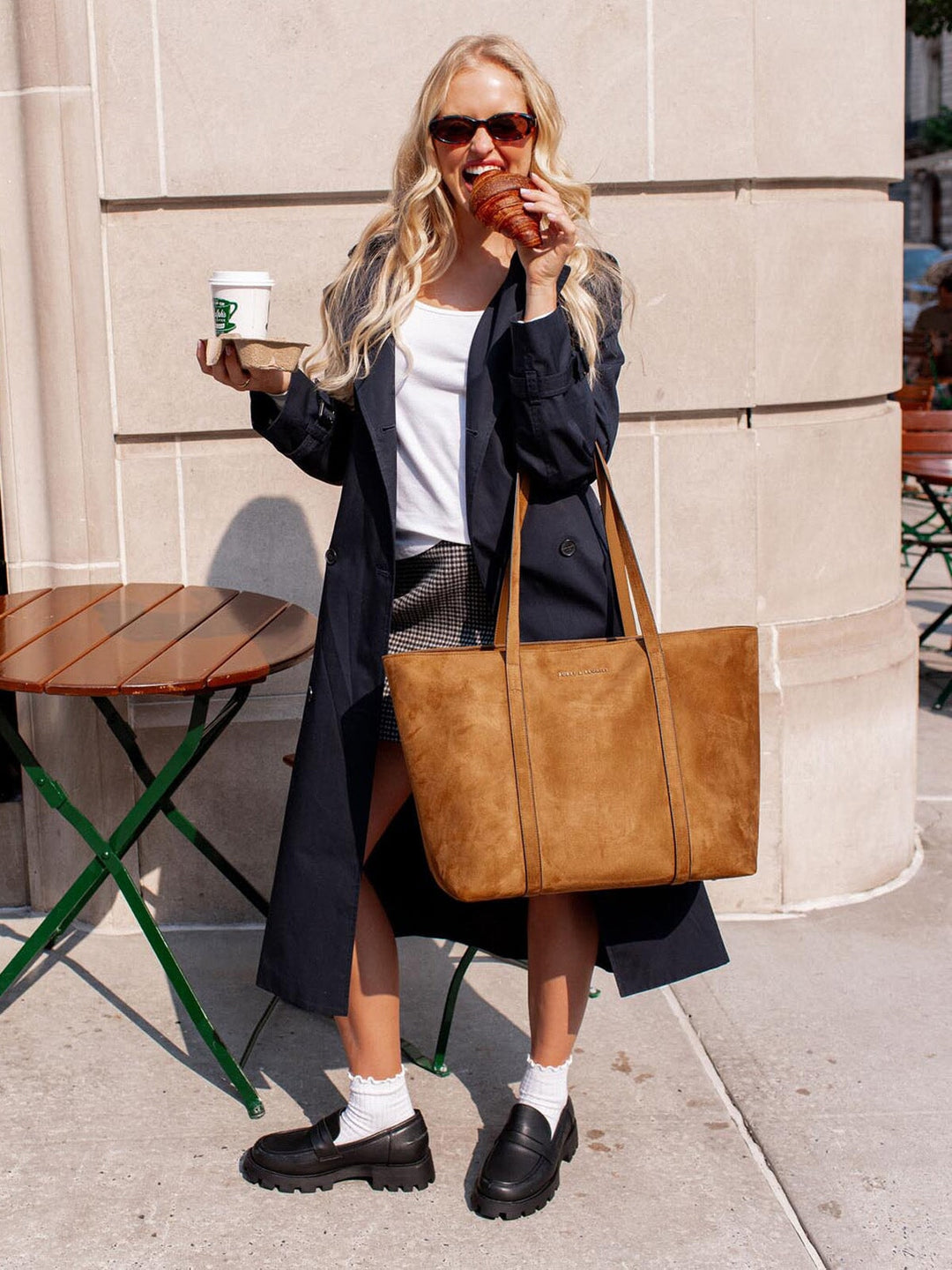 Woman holding a brown vegan suede tote bag, coffee cup, and croissant outdoors.