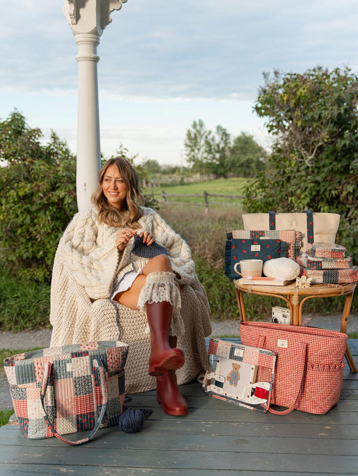 Poppy & Peonies Fallon Collection lifestyle image featuring Fallon knitting outdoors, surrounded by the Analog Tote in Strawberry Patchwork and Heart Berry Gingham, Strawberry Denim tote, coordinating project bags and craft accessories.