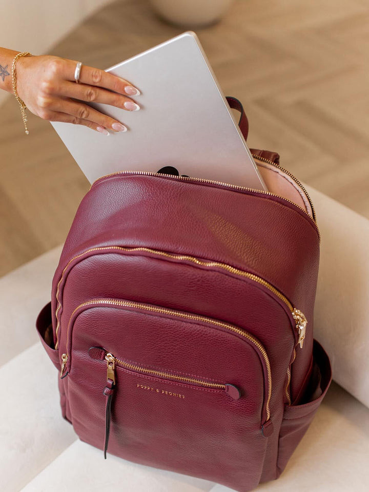 Person putting a laptop into a vegan leather backpack with gold zippers on a light wooden surface.