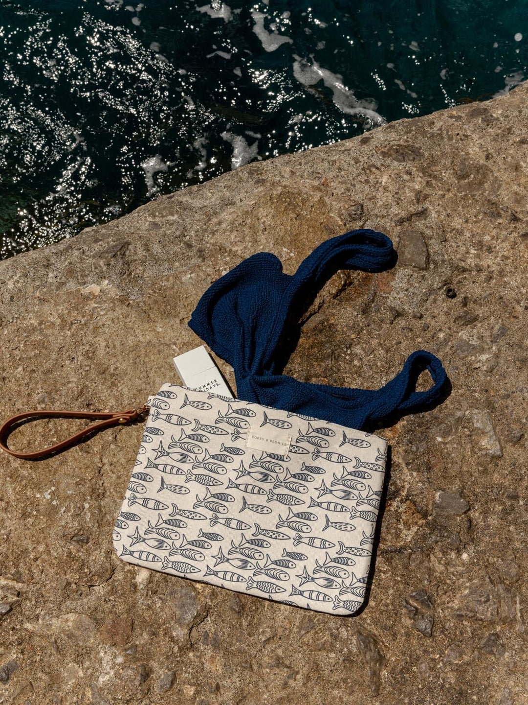 Small wet bag with a sardine pattern and blue drawstring on a stone surface near water.