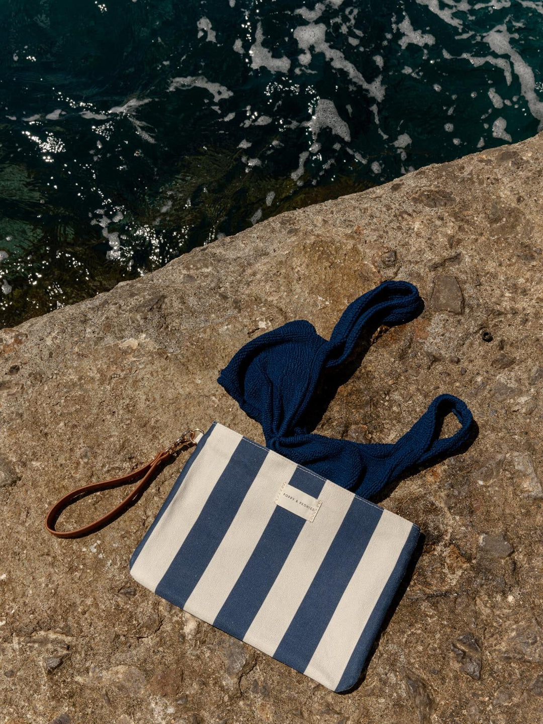 Blue and white striped wet bag with a blue bathing suit on a stone surface near water.