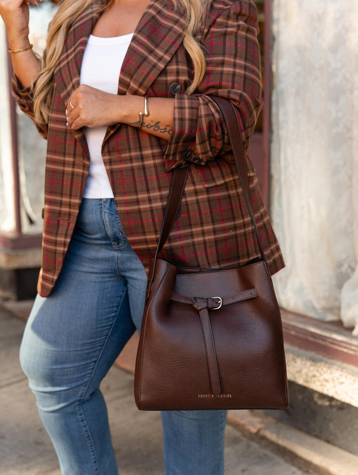 Poppy & Peonies The Knot Shoulder Bag in Espresso, close-up lifestyle view highlighting its rich brown exterior, decorative buckle, and shoulder strap against a plaid blazer.