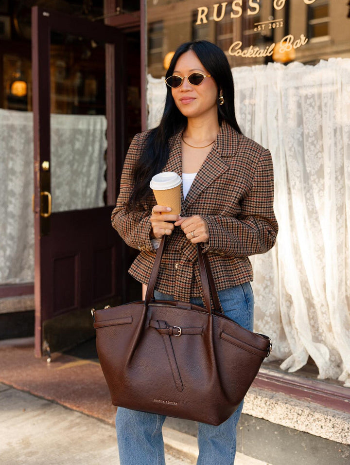 Poppy & Peonies The Knot Tote in Espresso, held by its long handles by a woman standing outside a café with a takeaway coffee, featuring its decorative buckle and spacious design.