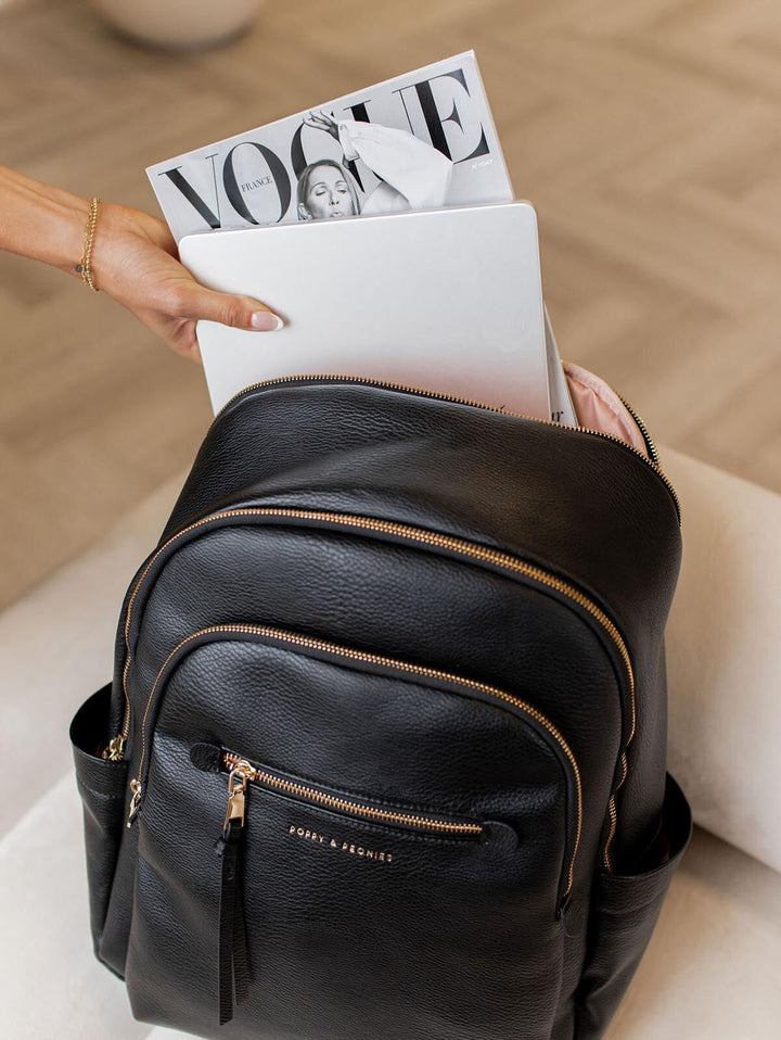 Black leather backpack with gold zippers holding a magazine and laptop on a neutral background