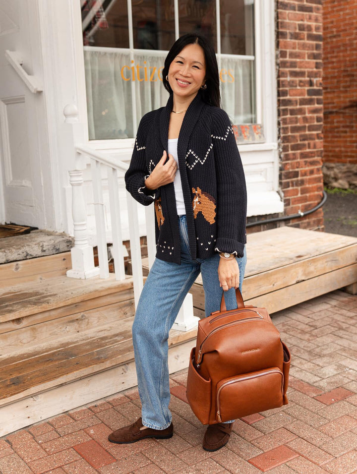 Poppy & Peonies The Cabin Backpack in Chestnut carried by its top handle by a woman in a navy cardigan and blue jeans outside a white storefront.