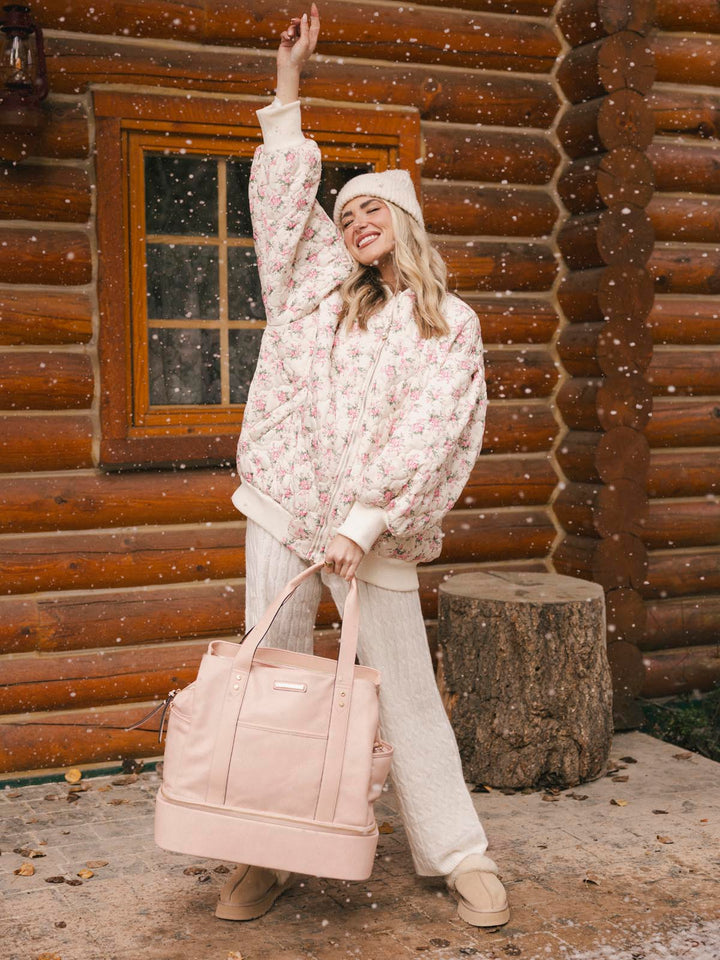 Woman in a floral sweater and white pants standing in front of a wooden cabin, holding a blush coloured vegan leather travel bag.