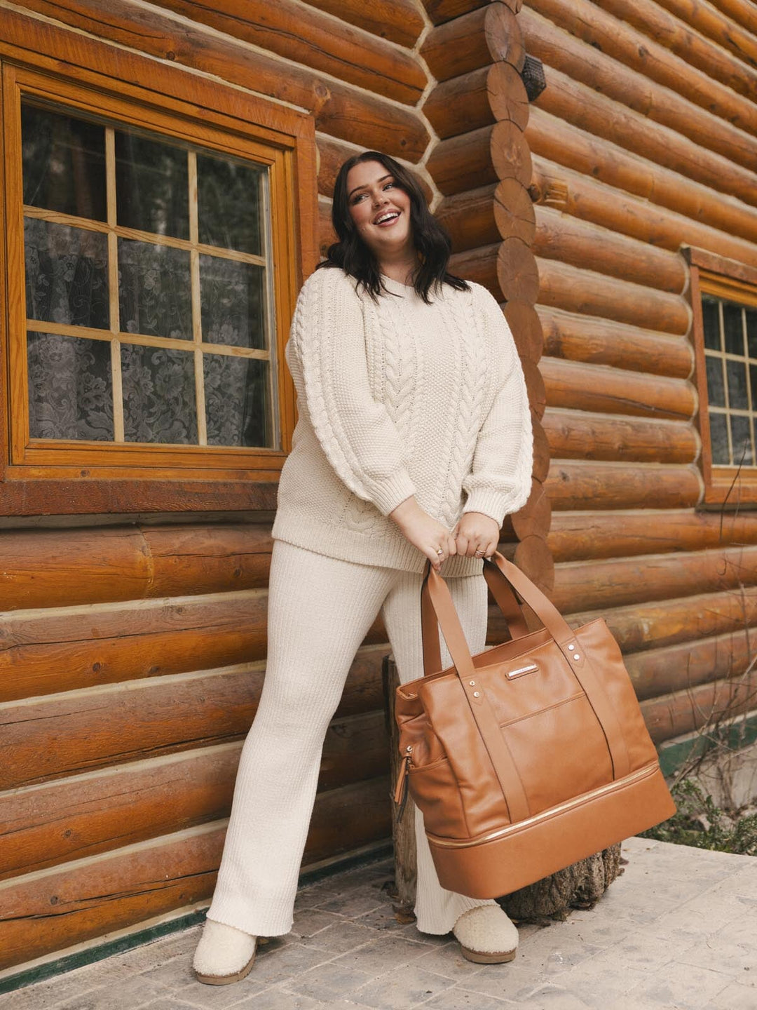 Woman holding a brown vegan leather bag in front of a wooden cabin