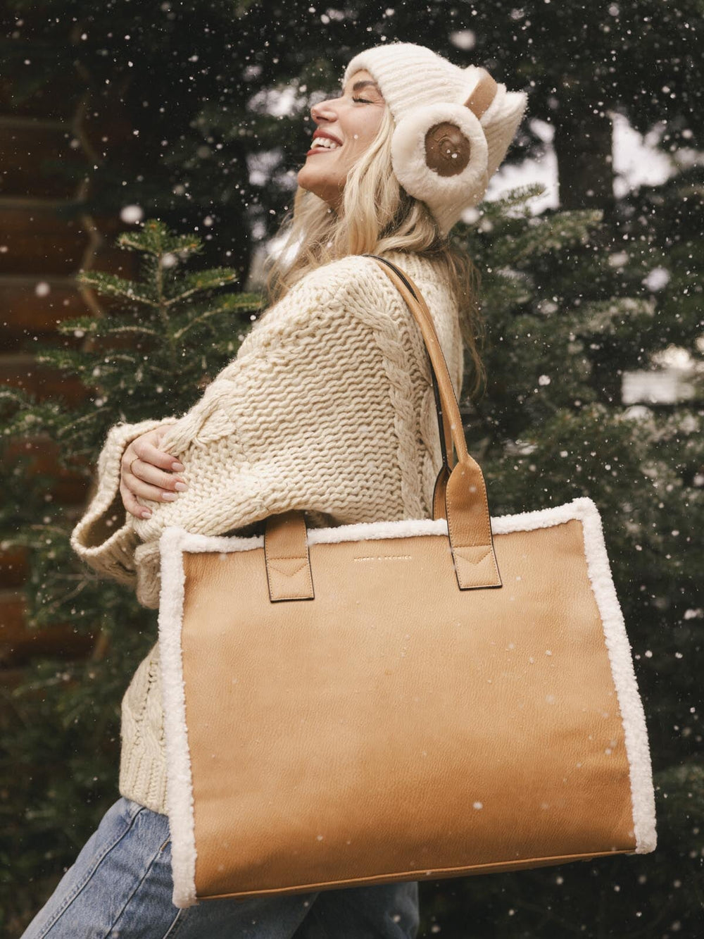 Person wearing a cream knit sweater and hat holding a brown vegan leather tote bag in a snowy setting.