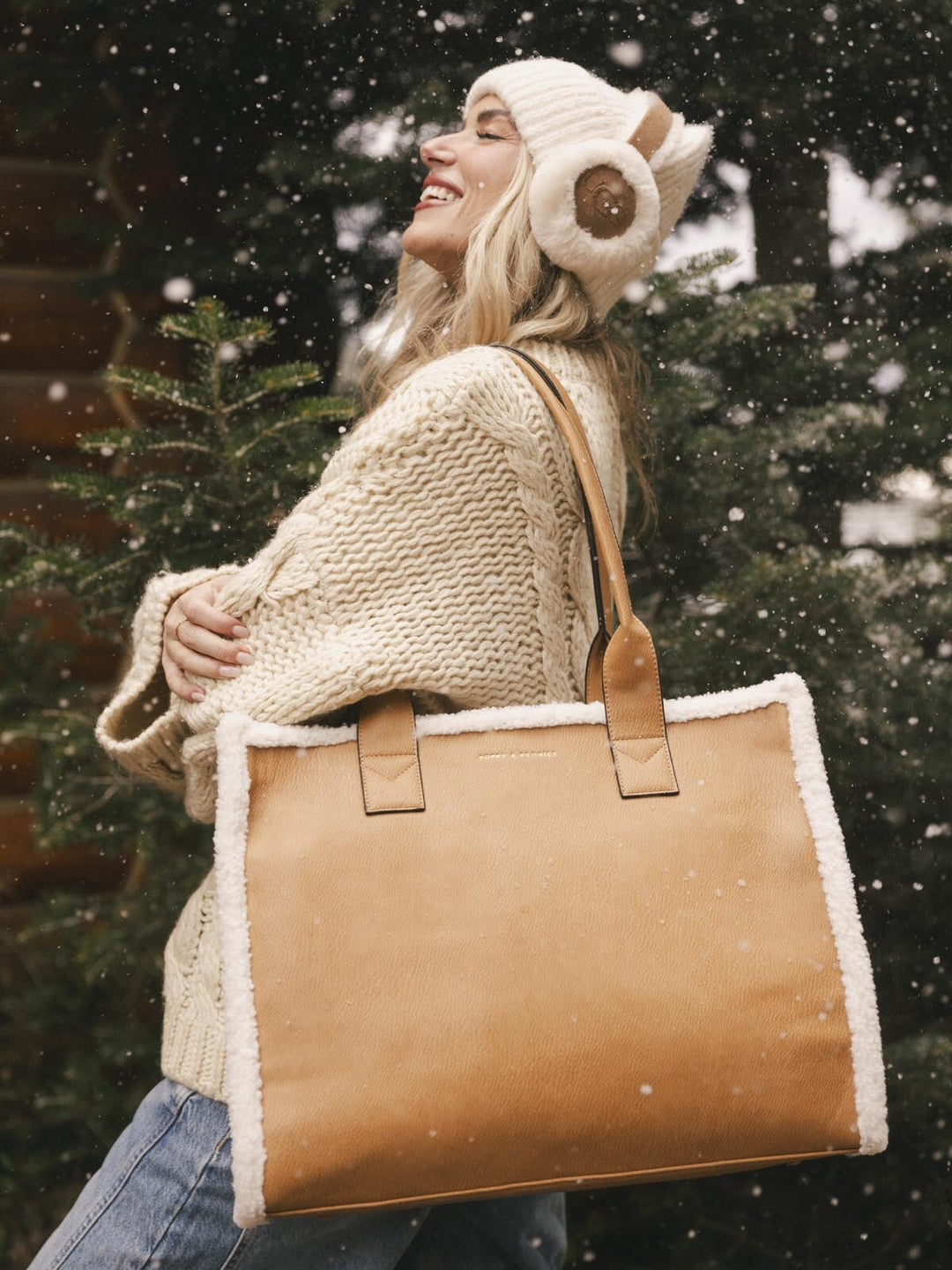 Person wearing a cream knit sweater and hat holding a brown vegan leather tote bag in a snowy setting.