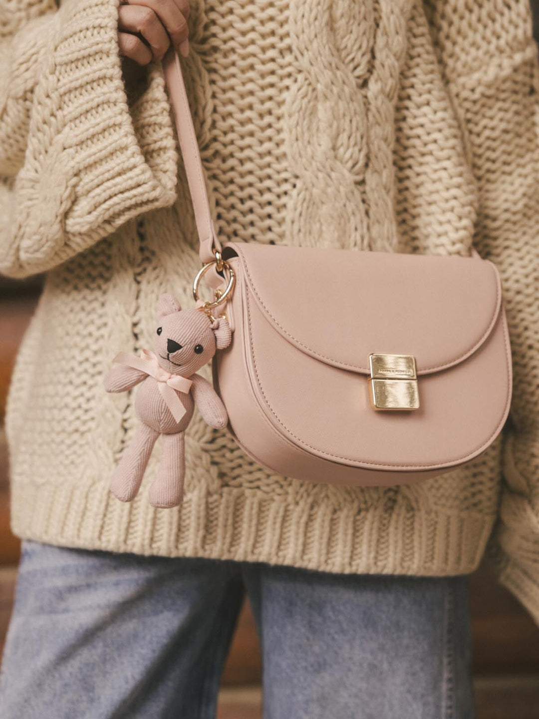 Pink crossbody bag with a teddy bear bag charm  held by a person wearing a beige knitted sweater.