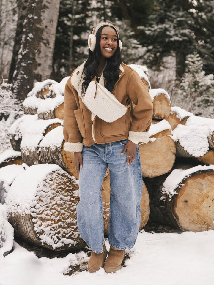 Person wearing a cream faux fur  crossbody bag standing in front of snow-covered logs.