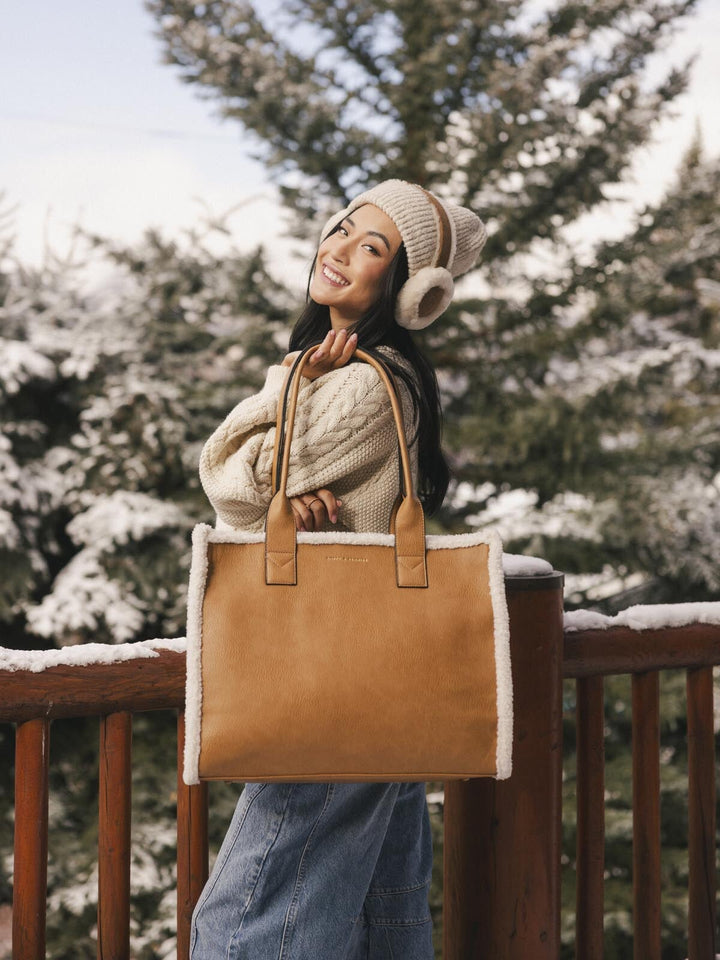 Person holding a brown vegan leather tote bag with a snow-covered pine tree background