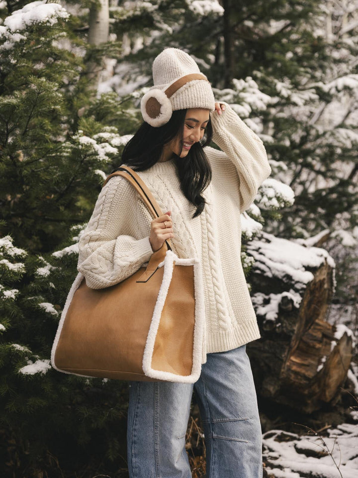 Woman in a snowy forest wearing a white knit sweater, blue jeans, and a brown vegan leather tote bag.