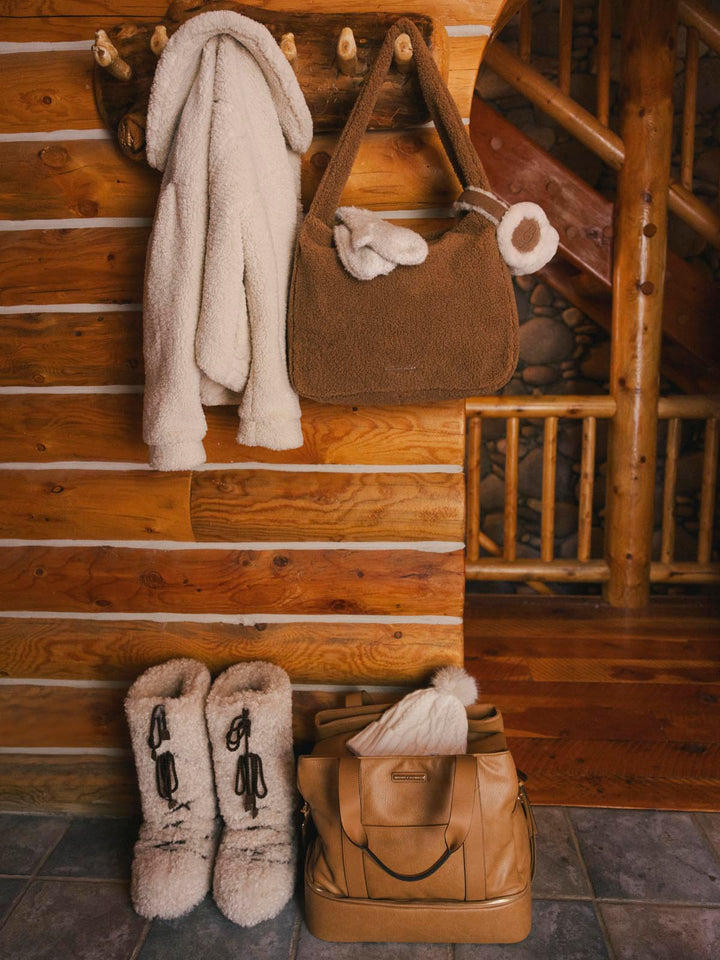 Brown faux fur bag, brown vegan leather travel bag, white moon boots, and beige coat in a cabin.