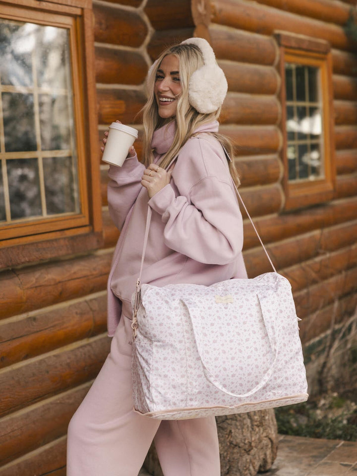 Woman in pink outfit with floral print duffel bag and ear muffs standing in front of a log cabin.