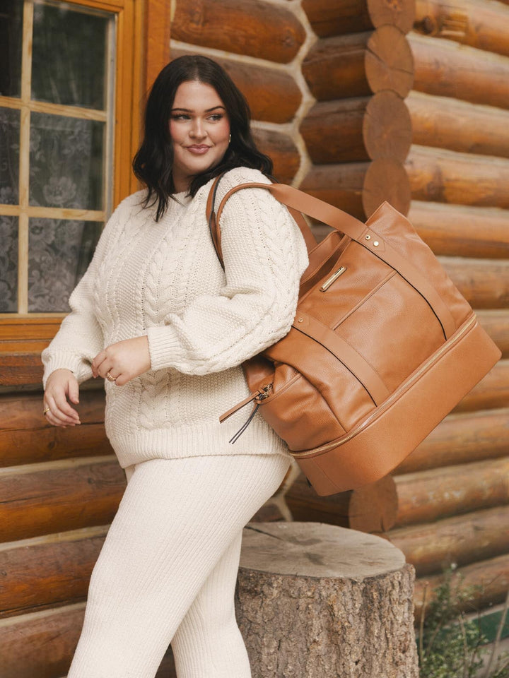 Woman in a white sweater with a brown vegan leather travel bag standing in front of a log cabin.
