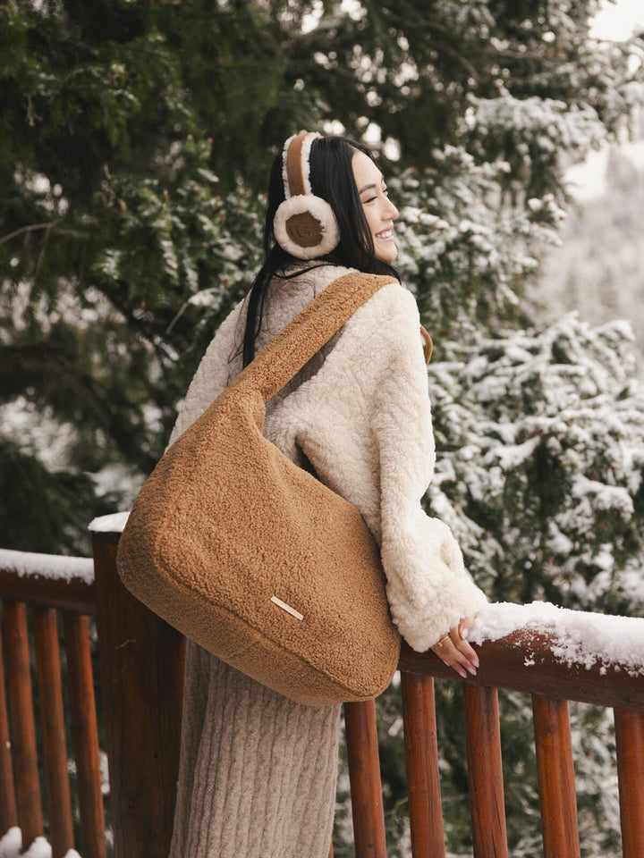 Person wearing a white coat and brown faux fur bag with ear muffs, standing on a snow-covered deck with trees in the background.