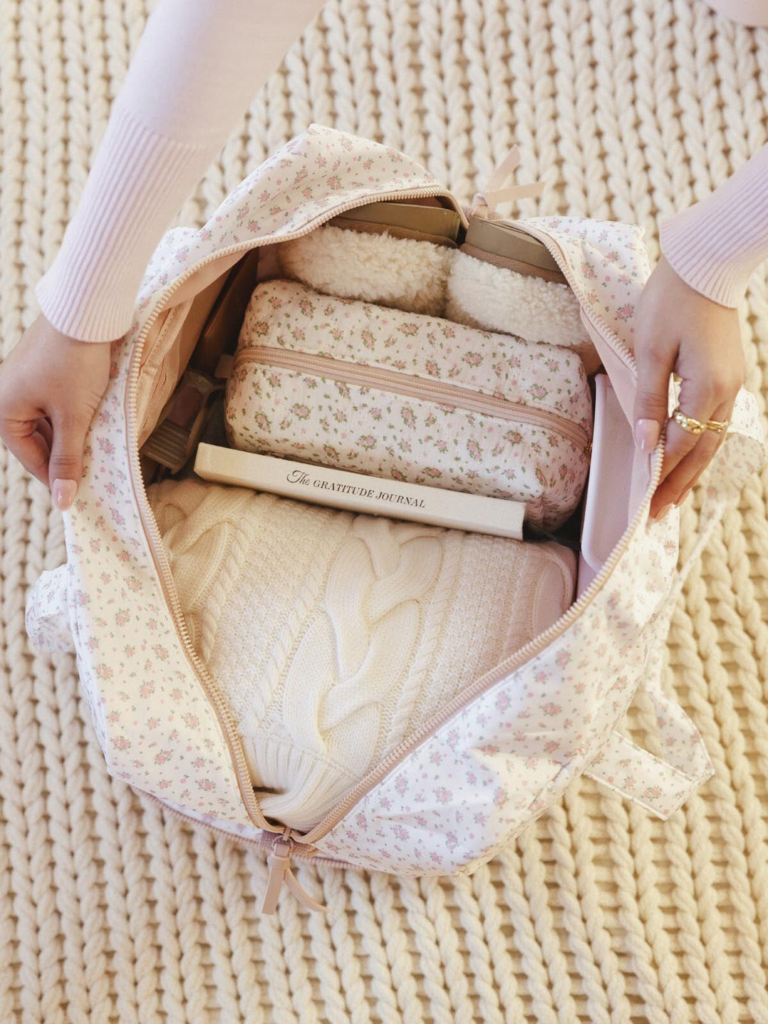 Person opening a floral-patterned travel bag filled with clothes and a book on a textured surface.