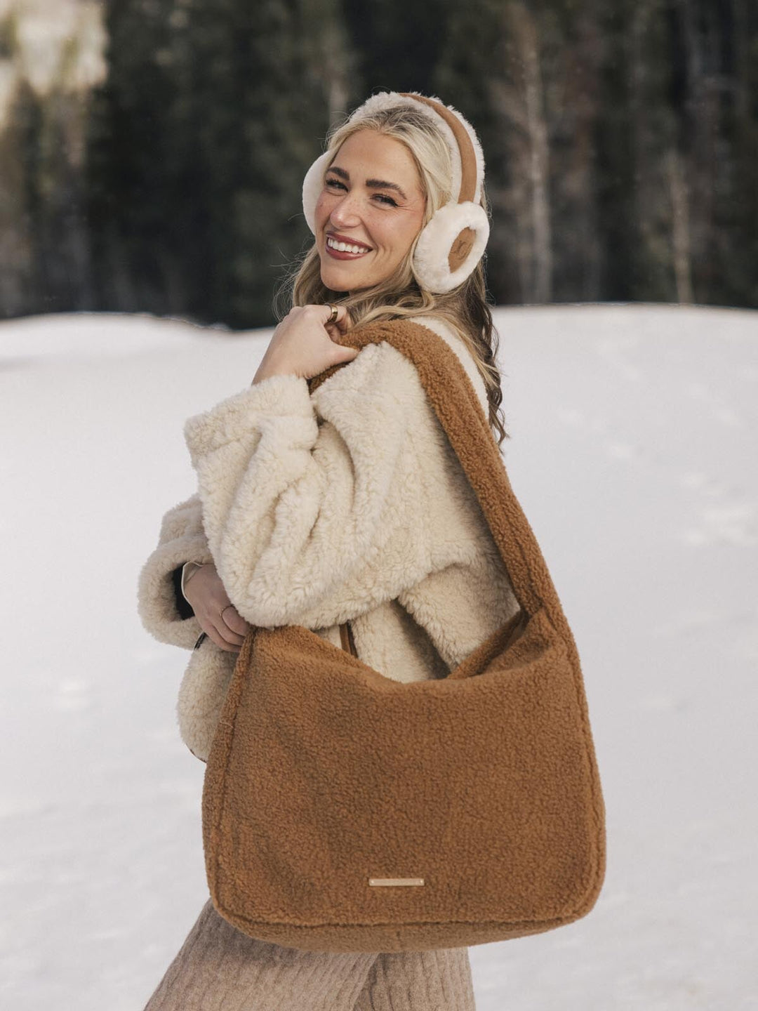 Woman in a snowy landscape wearing a brown faux fur bag, beige coat, and white ear muffs.