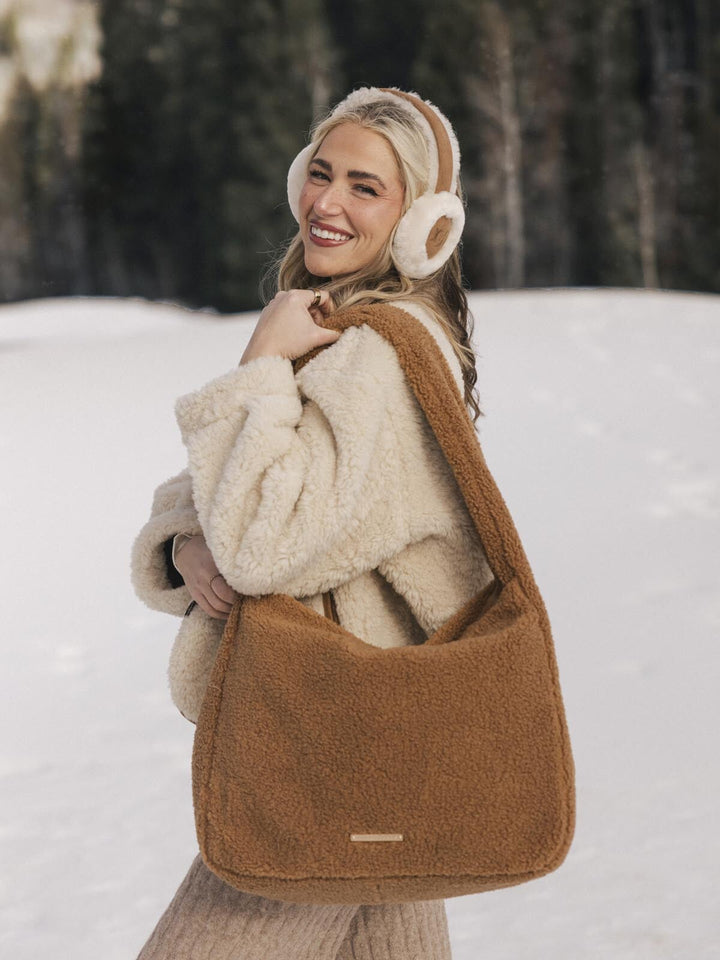 Woman in a snowy landscape wearing a brown faux fur bag, beige coat, and white ear muffs.