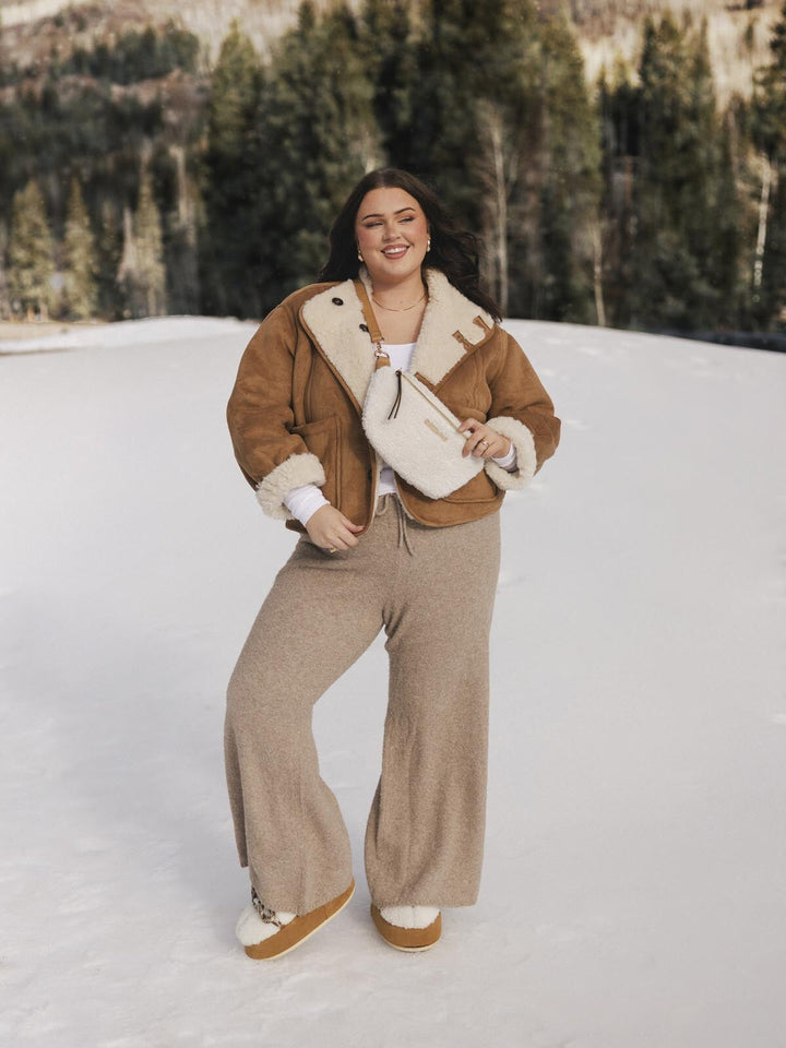 Person wearing a brown shearling jacket and cream faux fur crossbody bag standing in a snowy landscape with trees in the background.