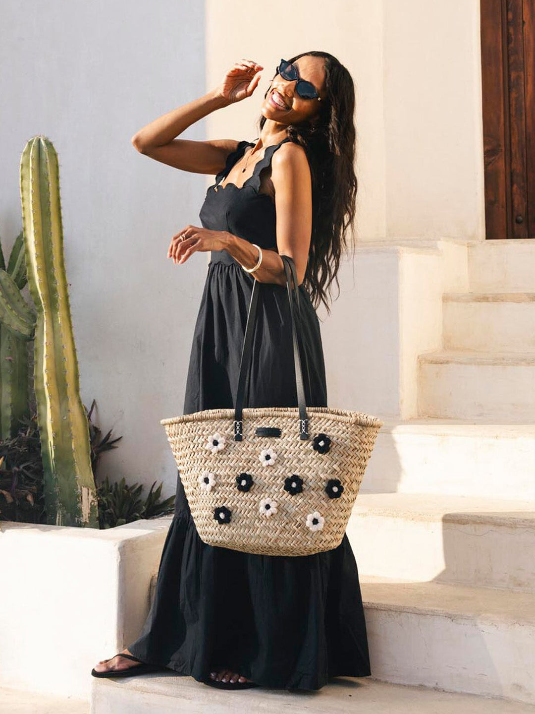 Woman in a black dress holding a straw bag with decorative elements, standing on steps with plants in the background.
