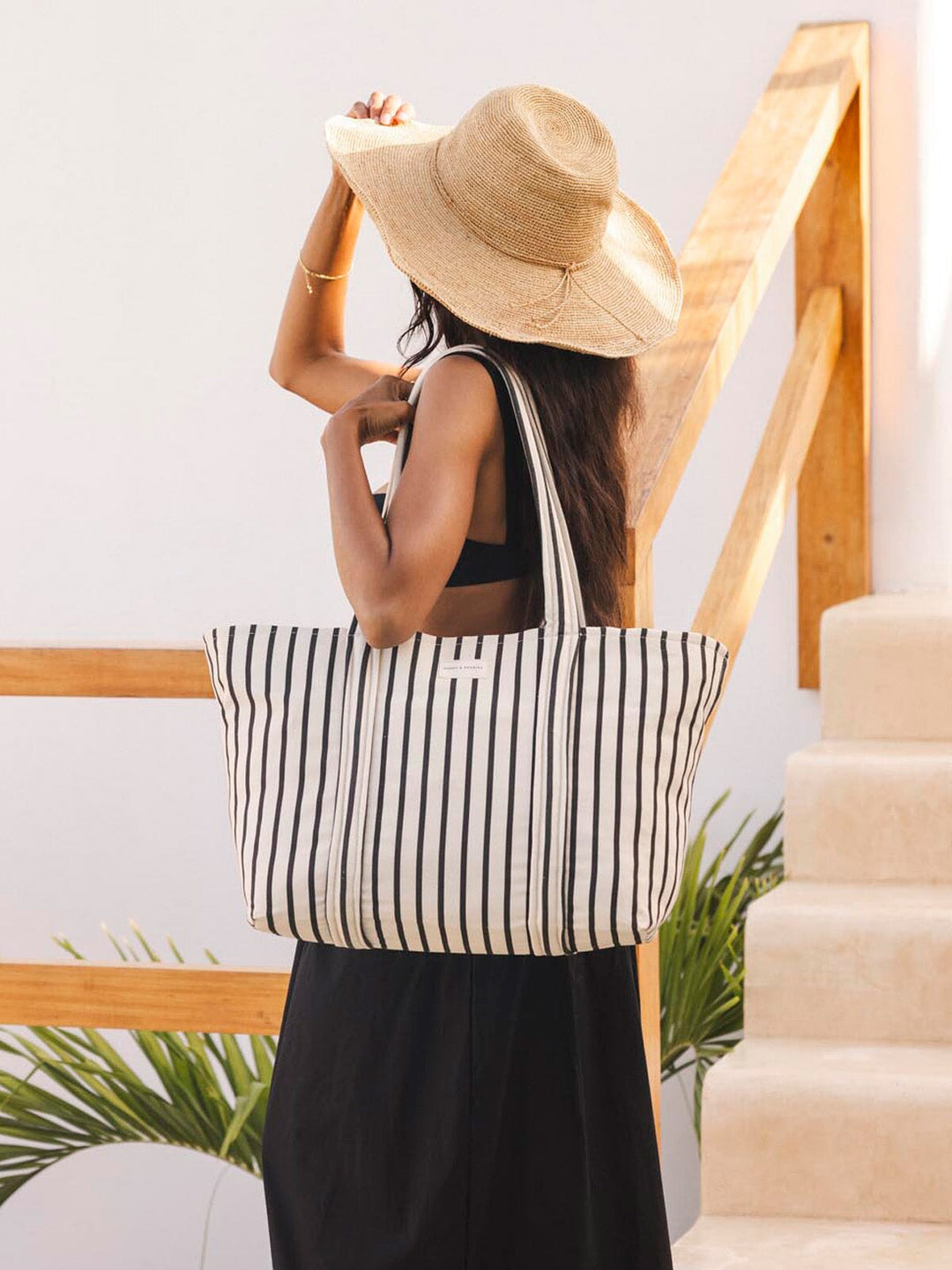 Woman holding a black and white striped tote bag with a straw hat, standing on a staircase.