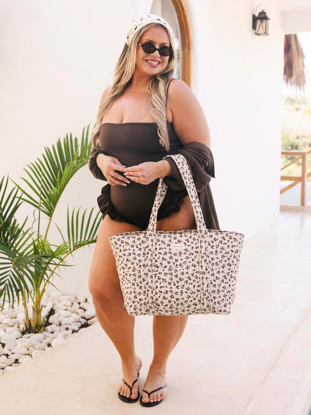 Woman in a brown swimsuit holding a floral patterned tote bag outdoors.