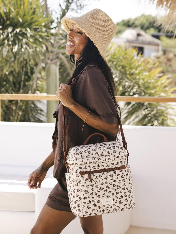 Woman with a floral-patterned lunch bag with brown straps and sun hat in a tropical setting
