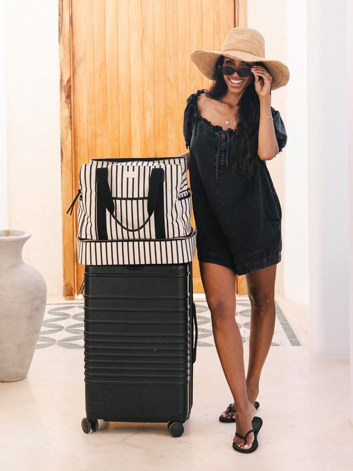 Woman in a black dress and hat standing next to a black suitcase with a striped travel bag on top.