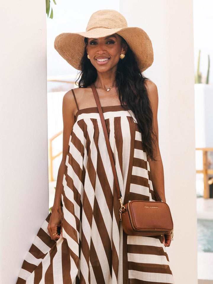 Woman wearing a brown and white striped dress with a wide-brimmed hat and carrying a brown vegan leather crossbody bag