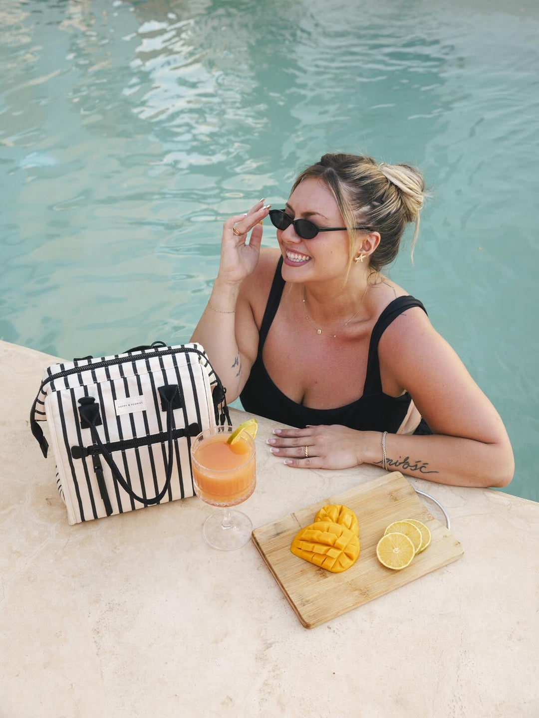 Woman by a pool with a black and white striped lunch bag, a drink and fruit, wearing sunglasses.