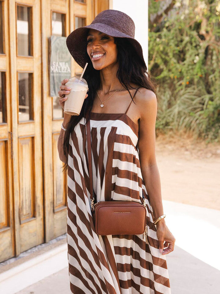 Woman in a brown and white striped dress holding a drink and wearing a brown vegan leather crossbody bag, standing outdoors.