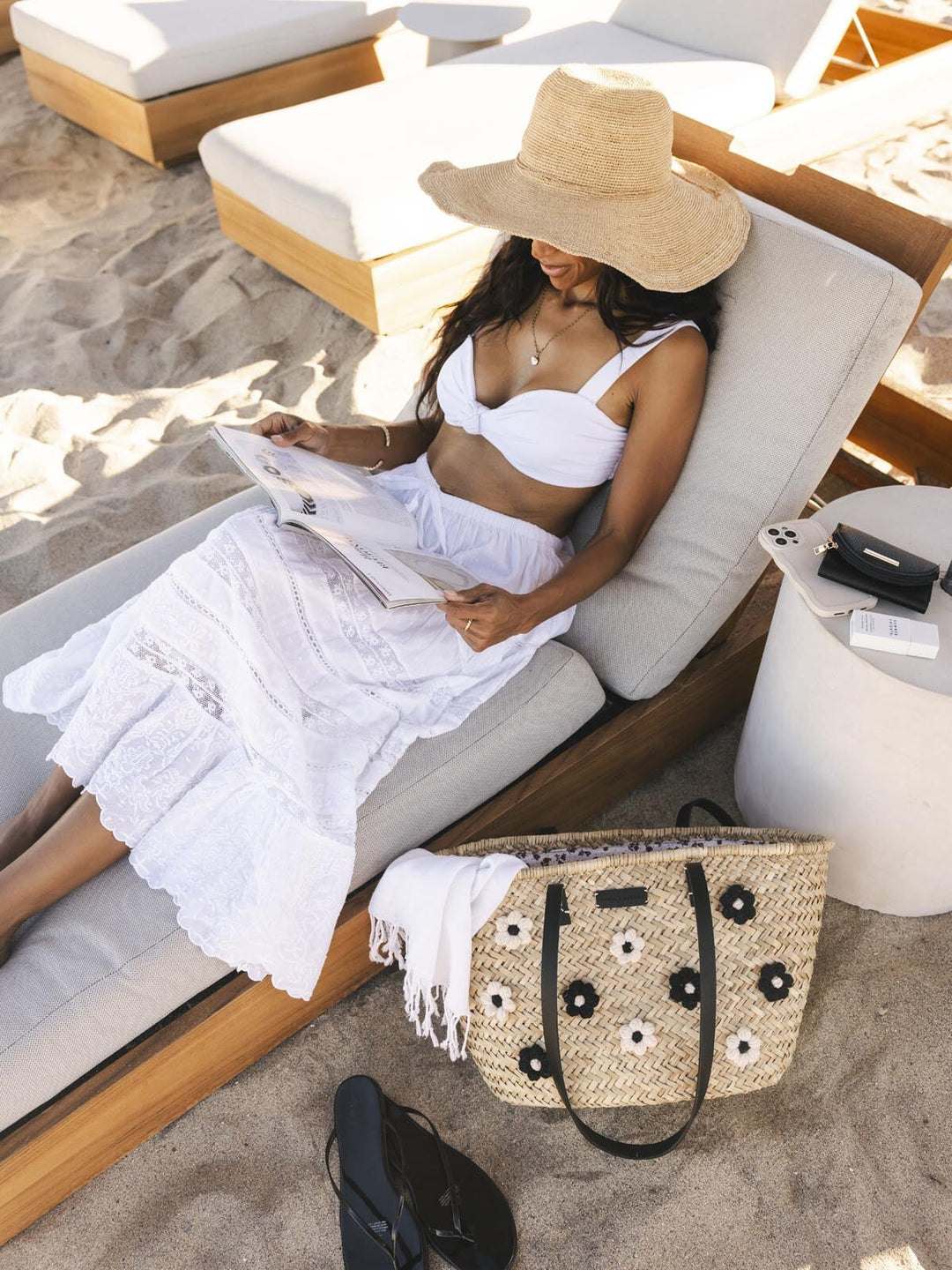 Woman in white dress and straw hat reading a magazine on a lounge chair with a straw handbag beside her.