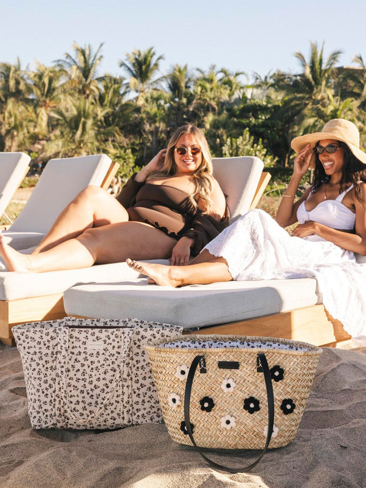 Two women lounging on beach chairs with a straw bag and a floral print tote bag in the foreground.